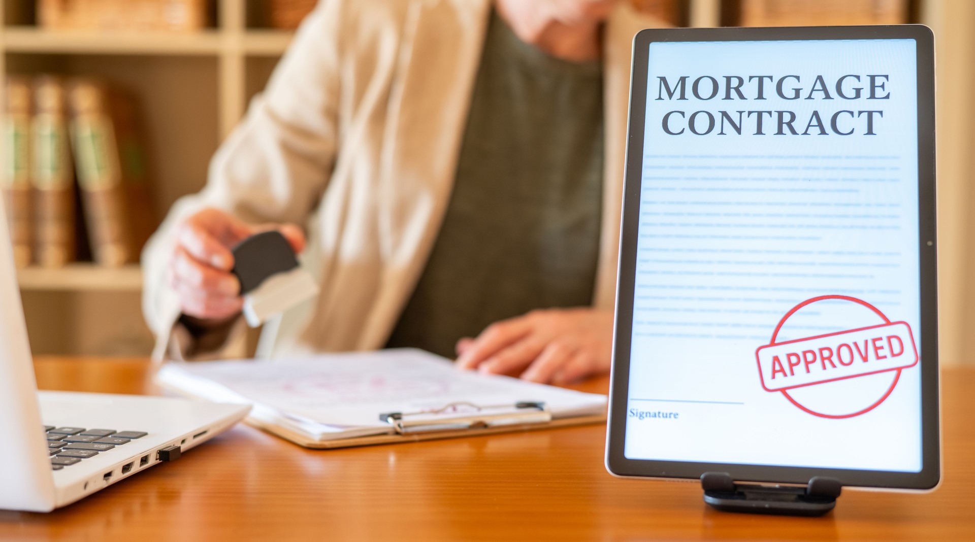 Person is stamping official paper documents while a digital mortgage contract with an approved stamp is displaying on a tablet screen, symbolizing loan approval and financial agreement
